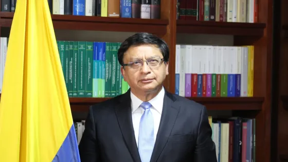 Fotografía de un magistrado de la Corte Constitucional de Colombia, de pie junto a la bandera nacional y frente a una biblioteca con textos jurídicos. La imagen acompaña la noticia sobre la ponencia que propone anular la reforma pensional por presuntas irregularidades en su trámite en la Cámara de Representantes.